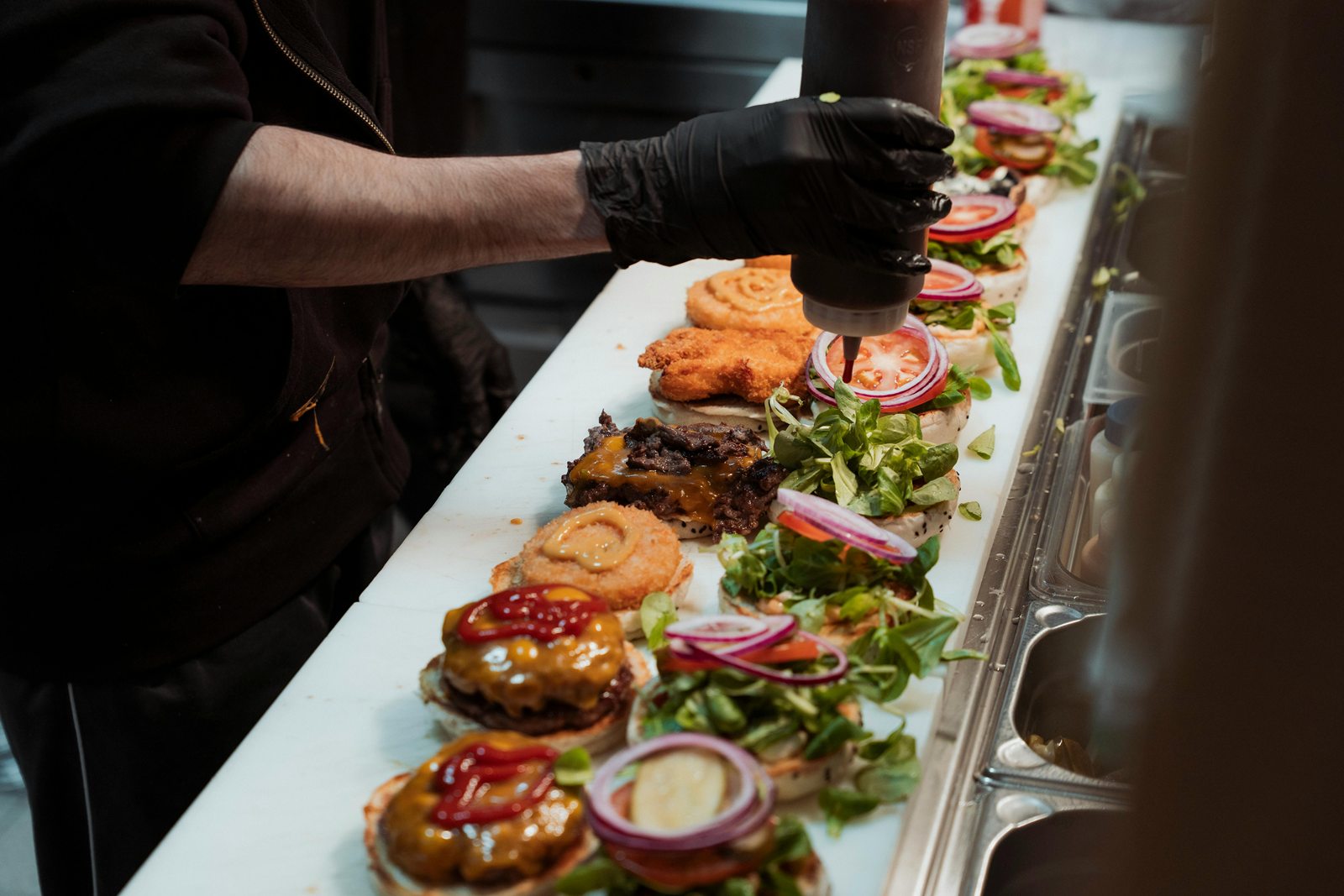 Een kok in zwarte handschoenen doet saus op een rij burgers met sla, tomaat en rode ui
